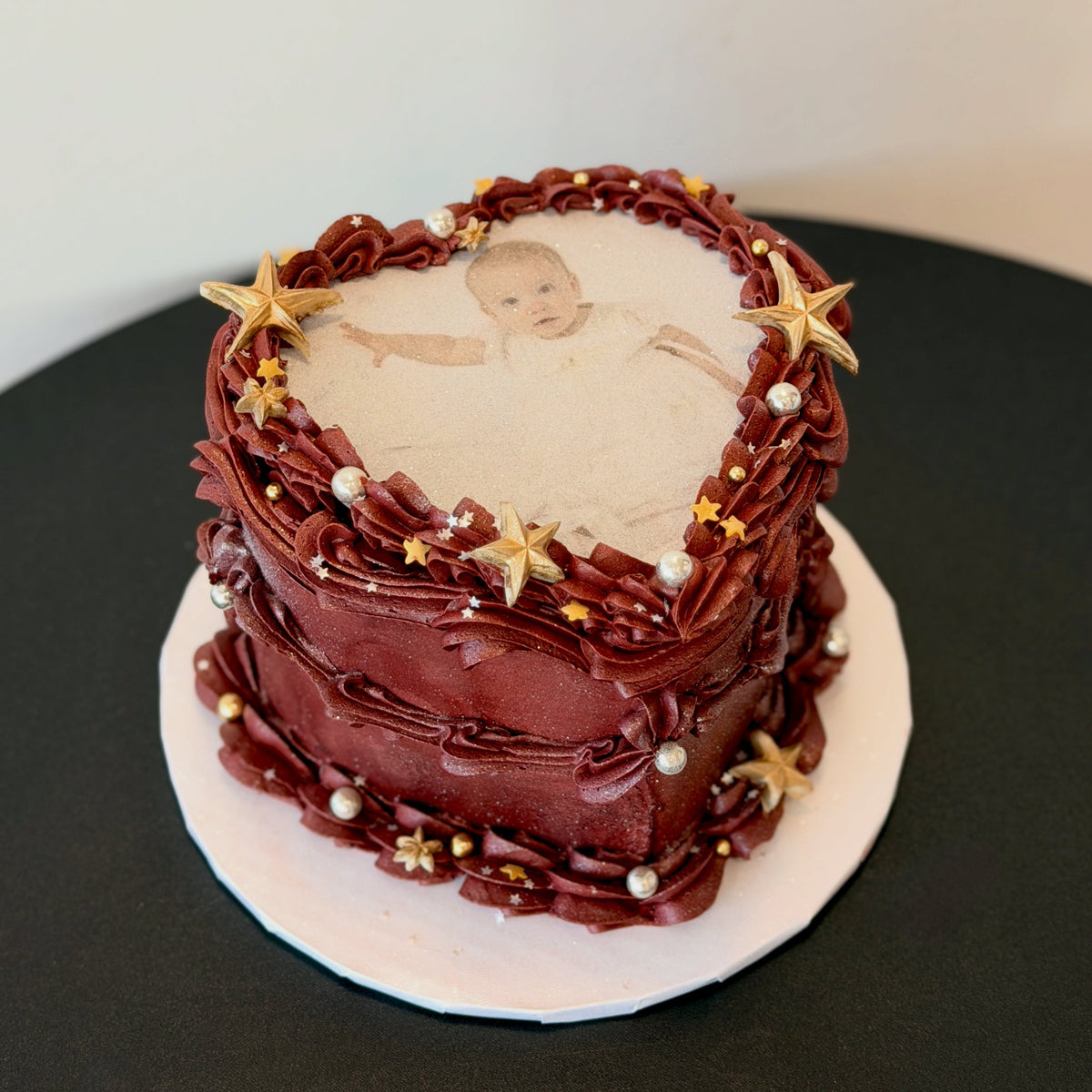 burgundy heart-shaped cake with a printed baby photo, gold stars and white pearls